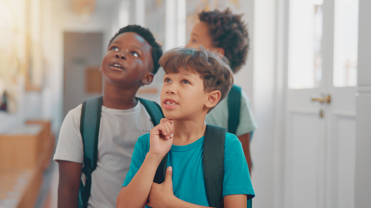 Three young students wearing backpacks look upward with curious expressions in a school hallway.