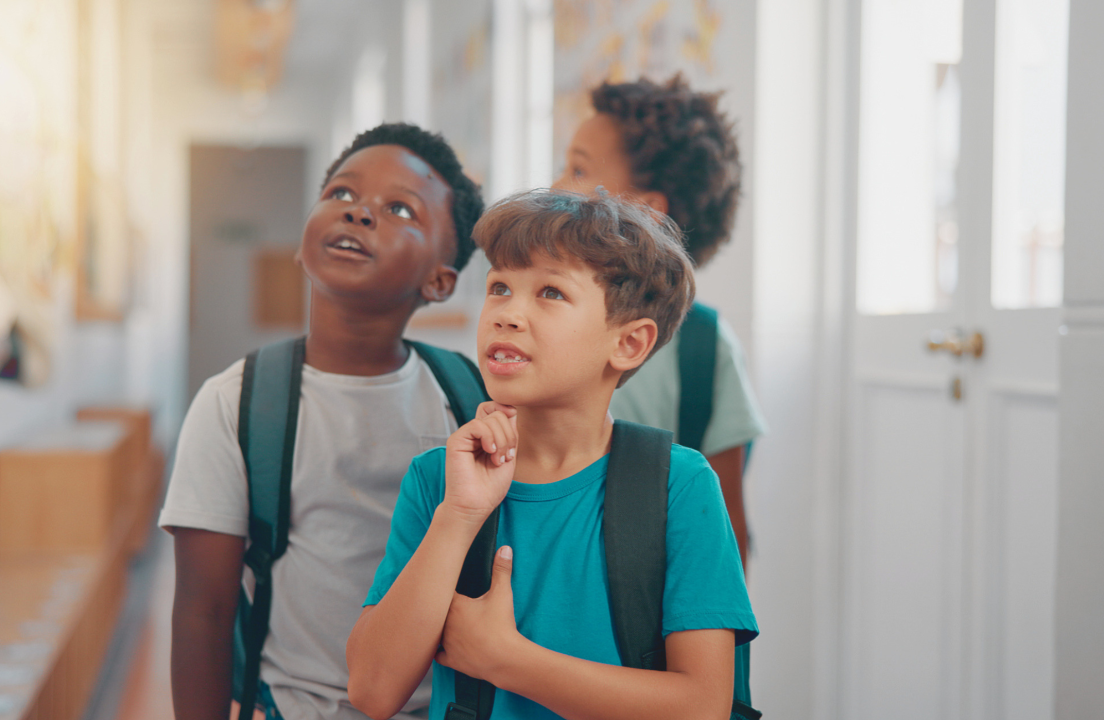 Three young students wearing backpacks look upward with curious expressions in a school hallway.