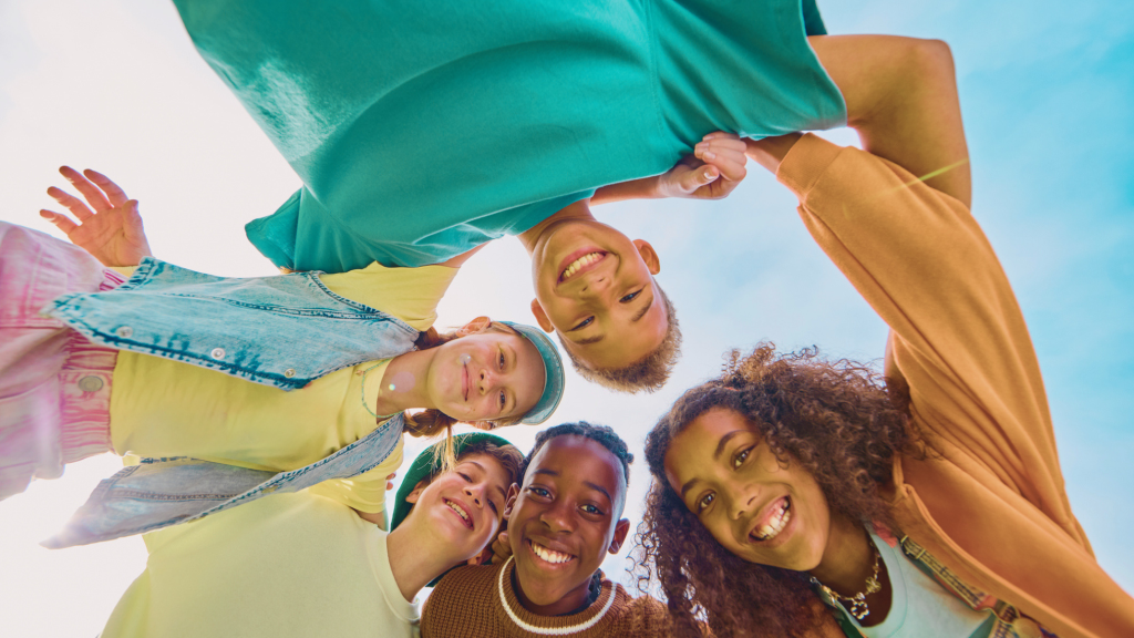 Five smiling teenagers huddle together and look down at the camera against a bright blue sky.