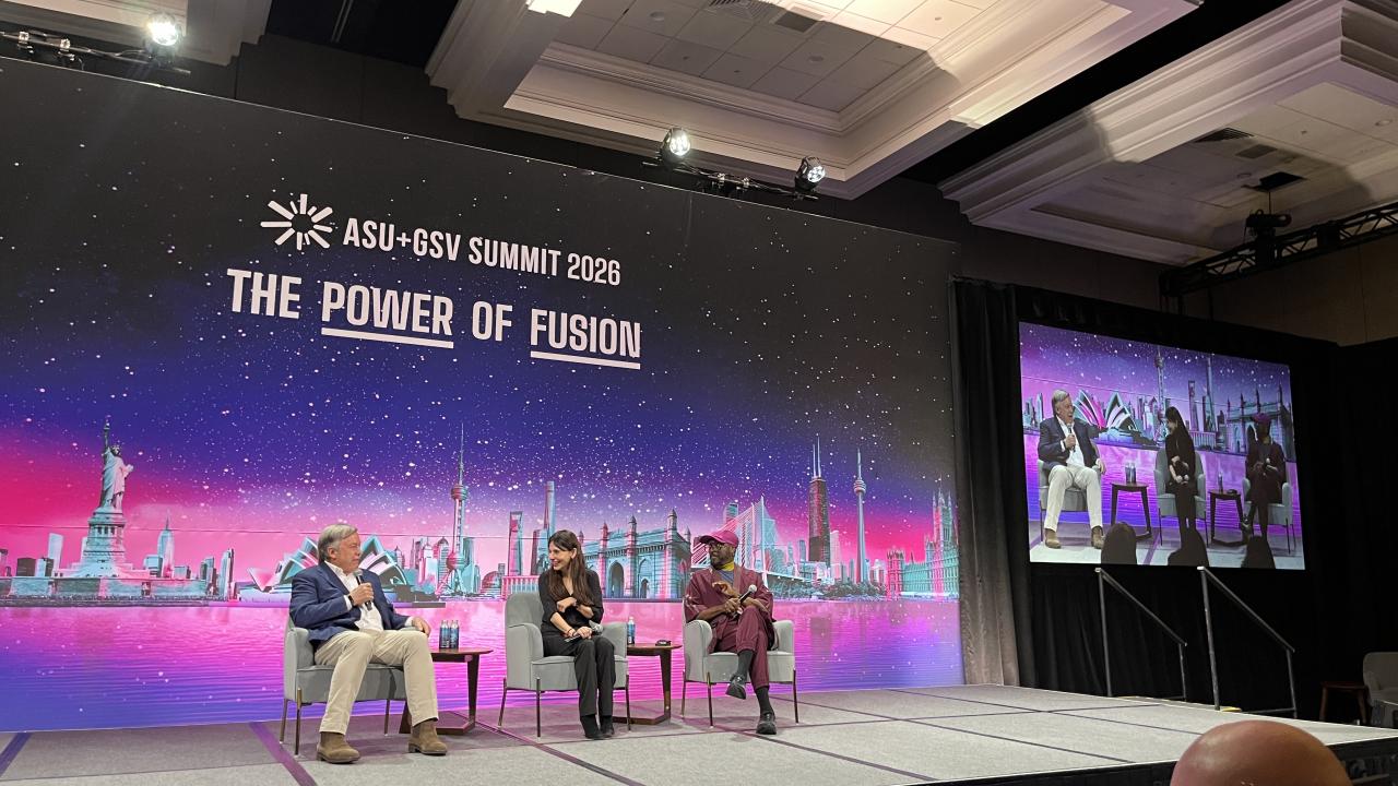 A panel discussion taking place on a stage at the ASU+GSV Summit 2026. The large backdrop reads “THE POWER OF FUSION” and features a colorful futuristic skyline with global landmarks against a starry purple and pink background. Three speakers sit in armchairs on stage, with one person speaking into a microphone while the others listen. A large screen to the right displays a live video feed of the discussion. Audience members are partially visible in the foreground.