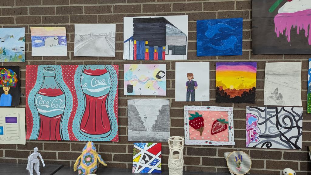 A student art display on a brick wall featuring paintings, drawings, and mixed media pieces, with ceramic vessels, crocheted figures, and embroidery arranged on a shelf below.