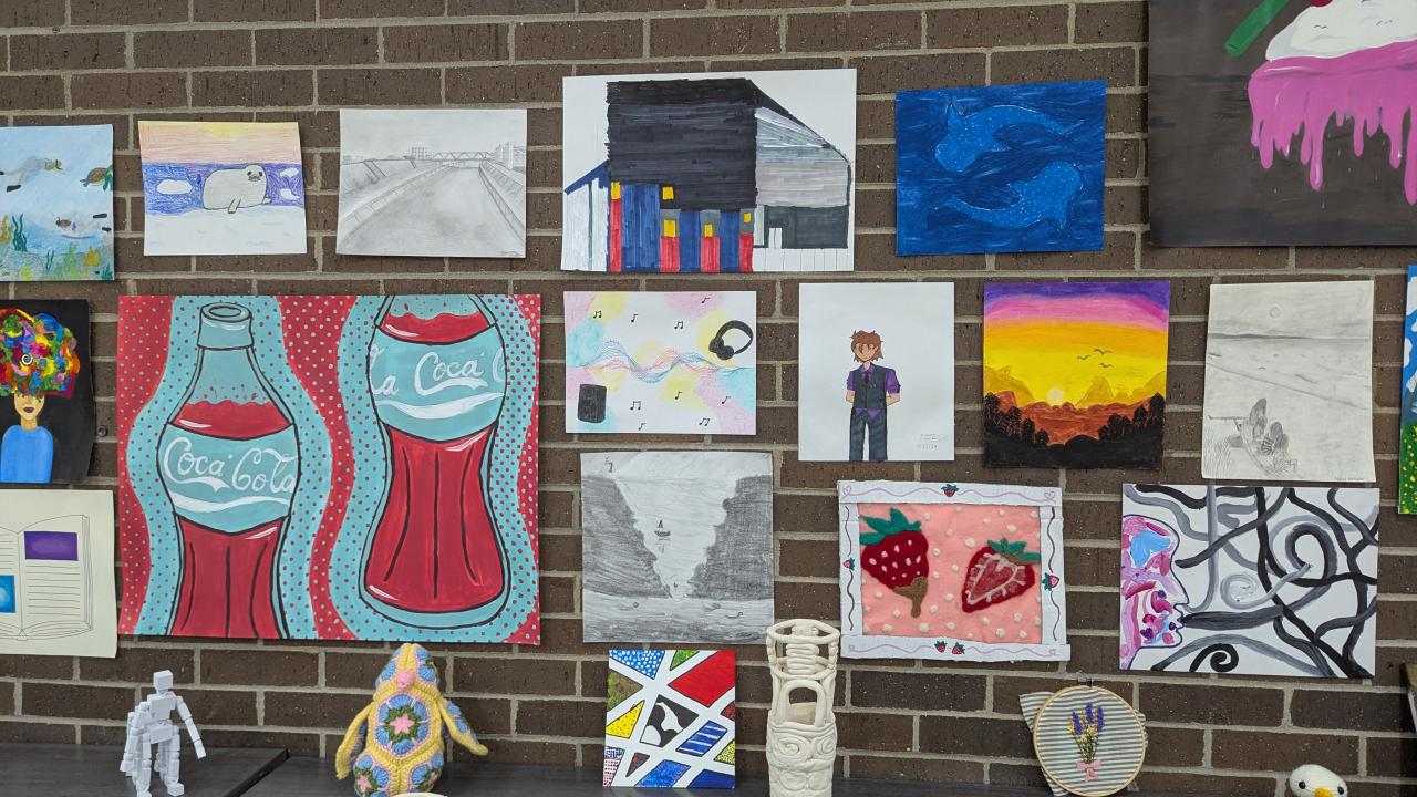A student art display on a brick wall featuring paintings, drawings, and mixed media pieces, with ceramic vessels, crocheted figures, and embroidery arranged on a shelf below.