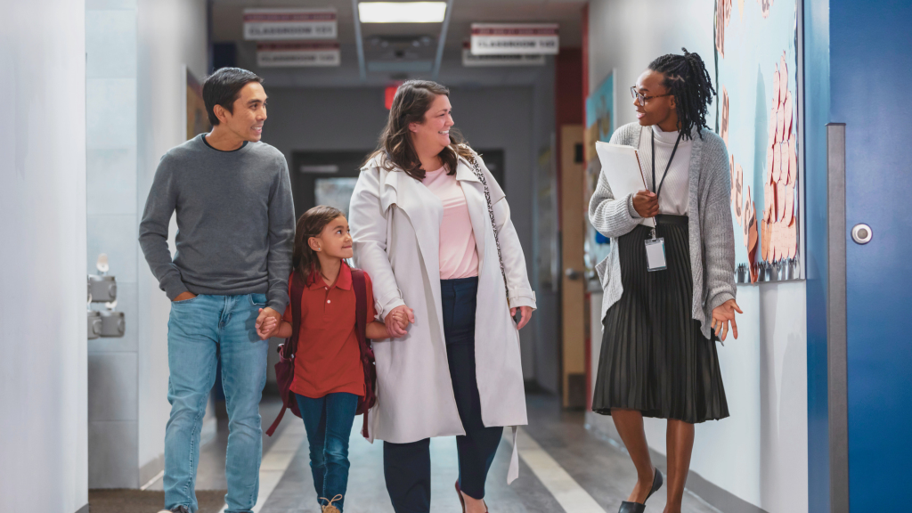 A teacher with a lanyard walks and talks with two parents and a young student in a school hallway.