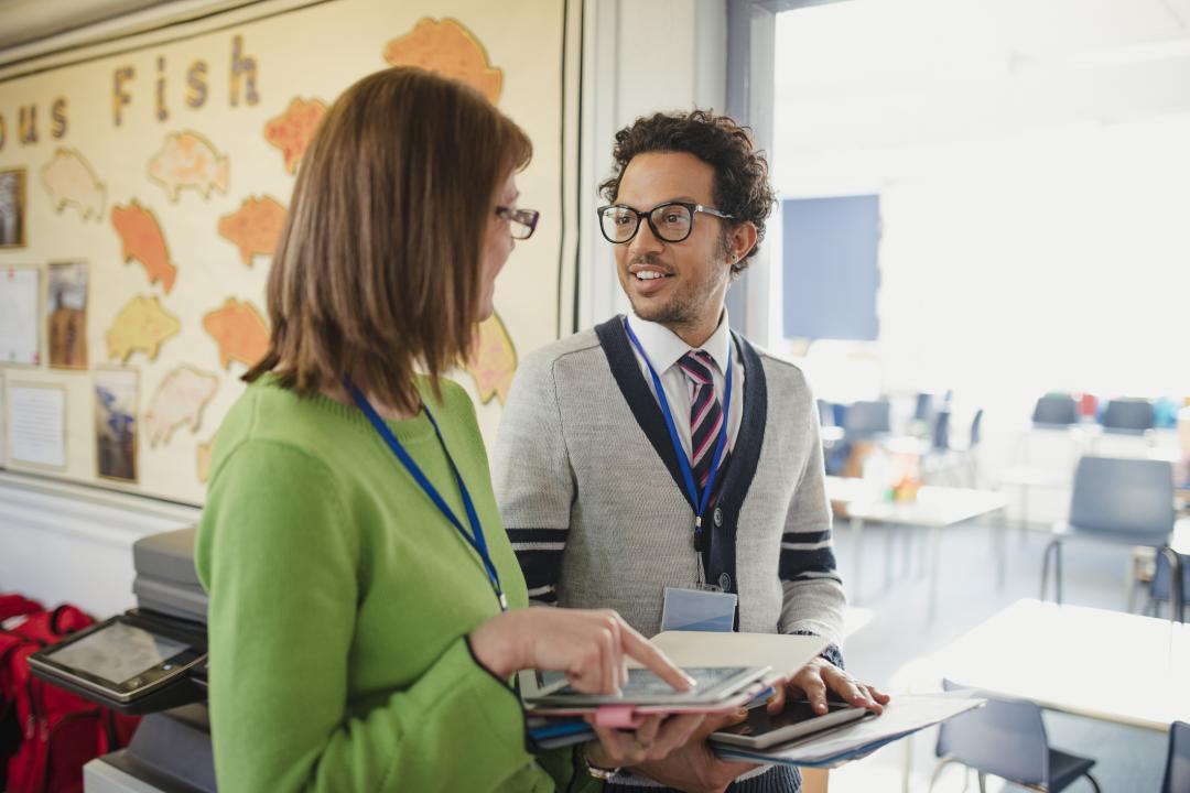 Two teachers wearing lanyards talk near a photocopier, one pointing to a tablet, in a classroom decorated with paper fish cutouts.