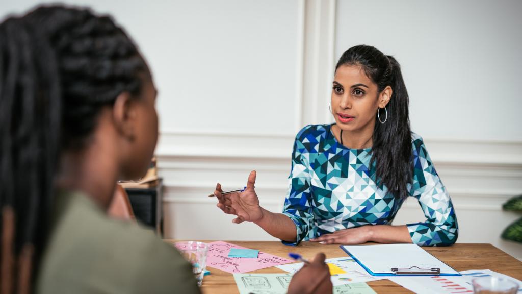 A woman gestures while discussing charts and sticky notes spread across a table during a meeting with a colleague.