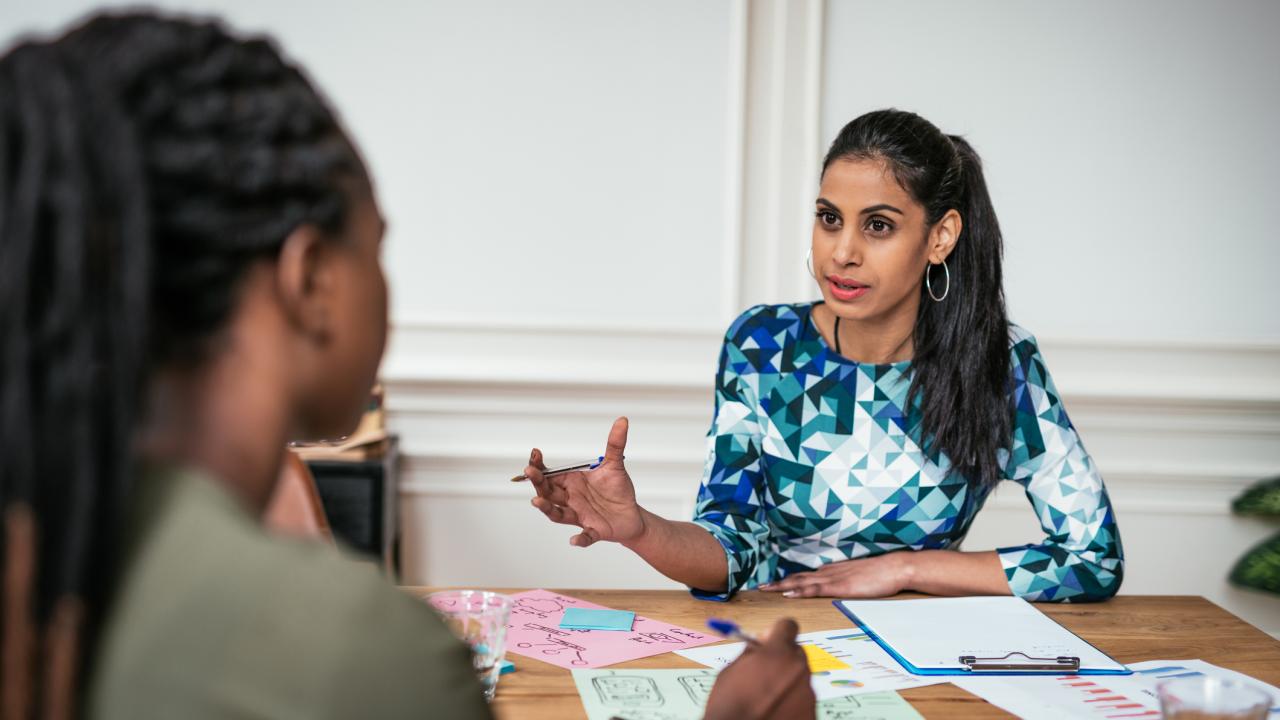 A woman gestures while discussing charts and sticky notes spread across a table during a meeting with a colleague.