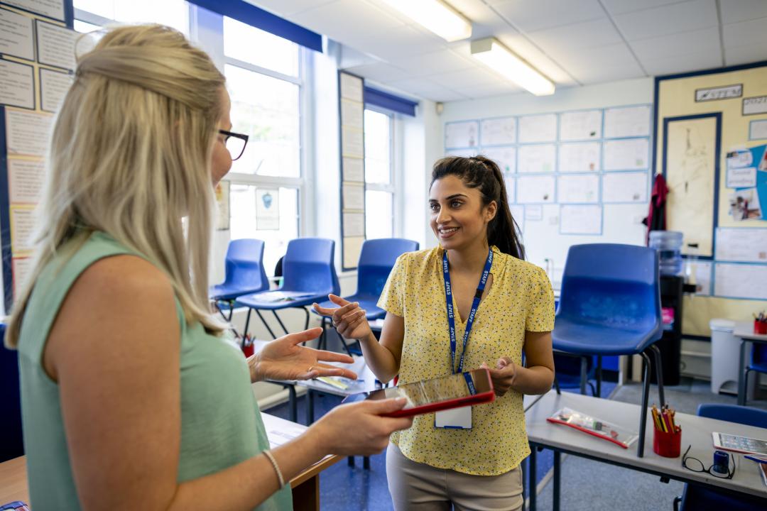 Two teachers chat and gesture while talking in a classroom, one holding a tablet, with empty chairs and a bulletin board in the background.