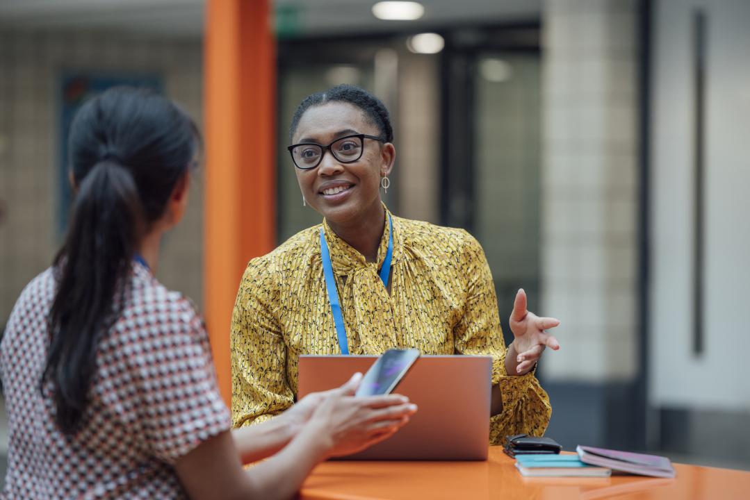 A smiling woman gestures while talking with another person seated at a table with a laptop, phone, and notebooks.