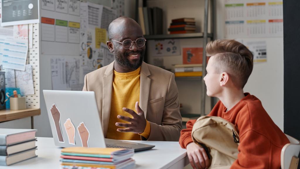 A teacher smiles and talks with a student sitting across a desk, with a laptop and stack of books between them in an office decorated with charts and calendars.