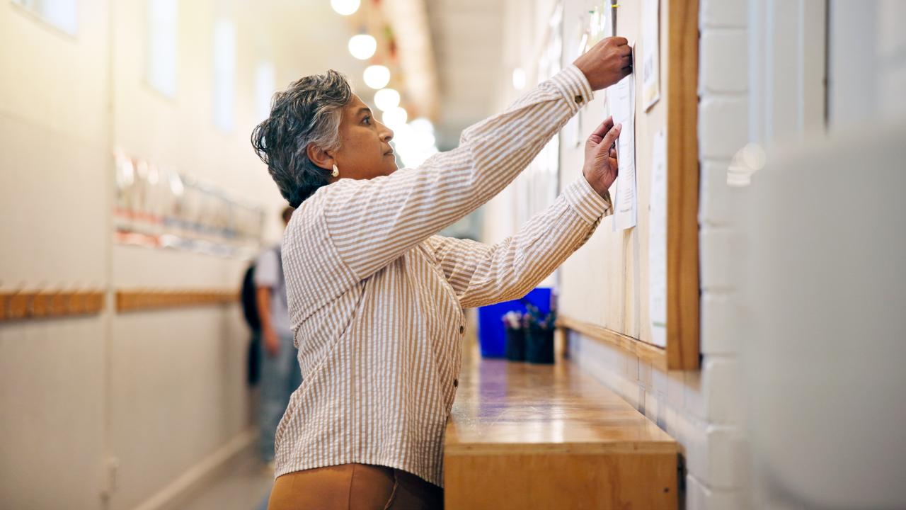A woman pins papers to a bulletin board in a school hallway.