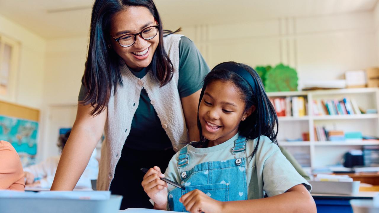 A teacher smiles while helping a student write in a notebook at her desk in a classroom with bookshelves and a world map.