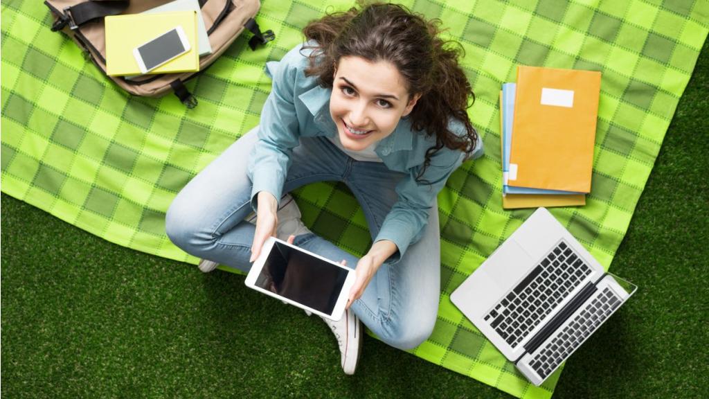 Young smiling student relaxing outdoors, she is sitting on the grass and using a laptop, summer camp concept