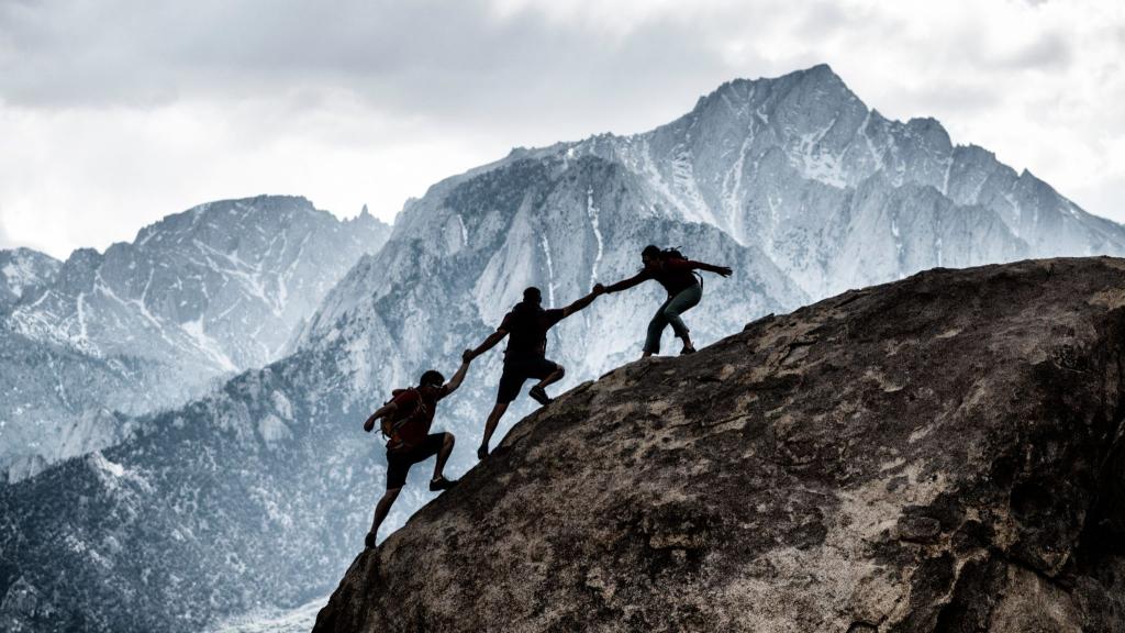3 men climbing a mountain. All three are helping eachother climb.