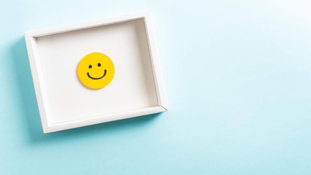 Concept of well-being, well done, feedback, employee recognition award. Happy yellow smiling emoticon face frame hanging on blue background with right empty space for text.