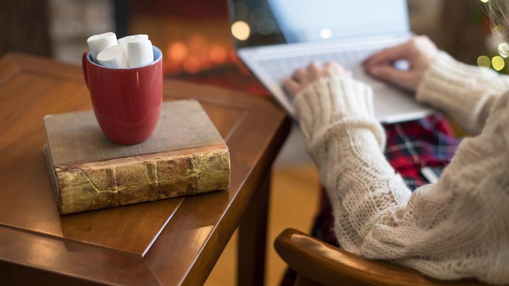 Woman hand with laptop, cup of hot cocoa and marshmallow on book on wooden table near christmas tree and fireplace.