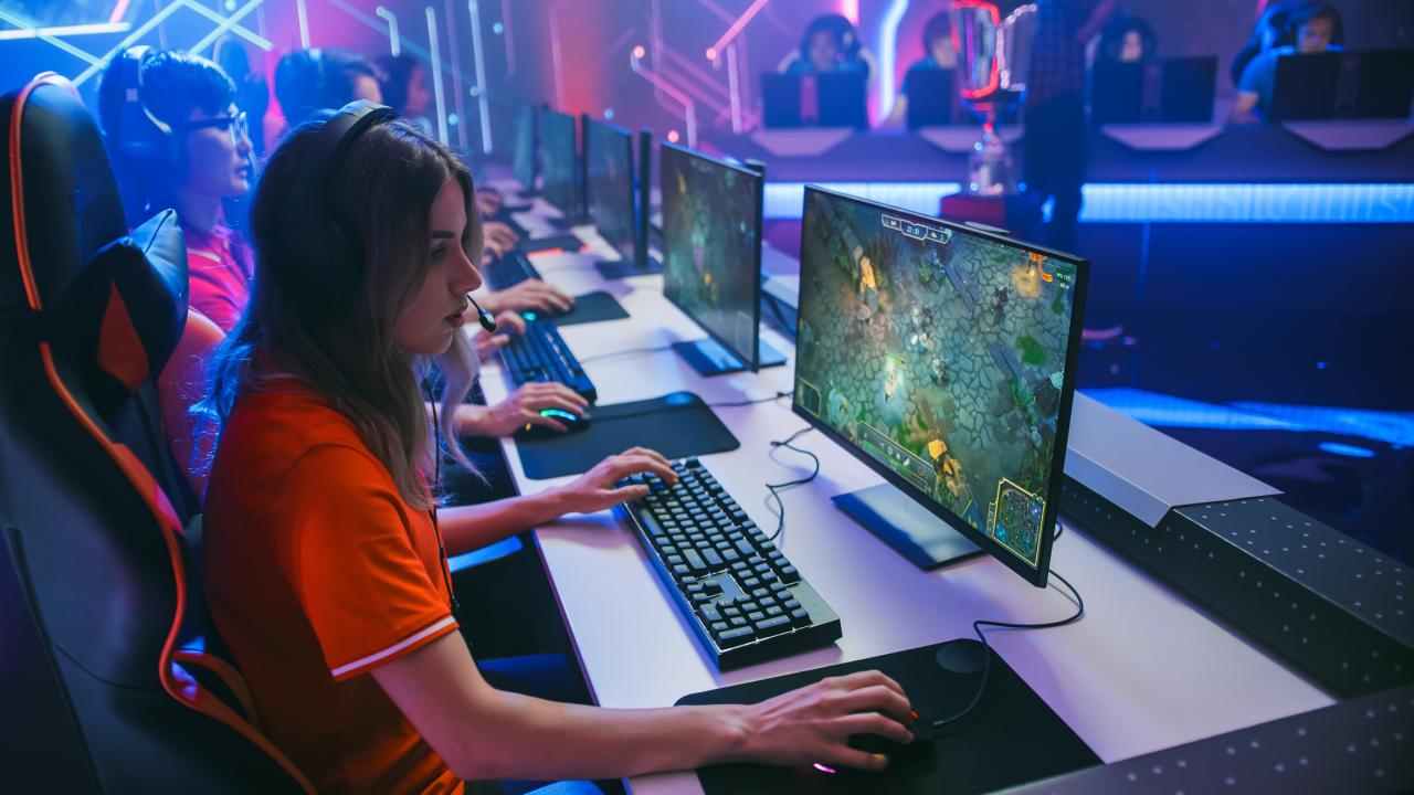 Dark room with glowing lights. 8 people are looking at computers and playing a game. They wear headsets and are focusing on the screen while typing on keyboards.
