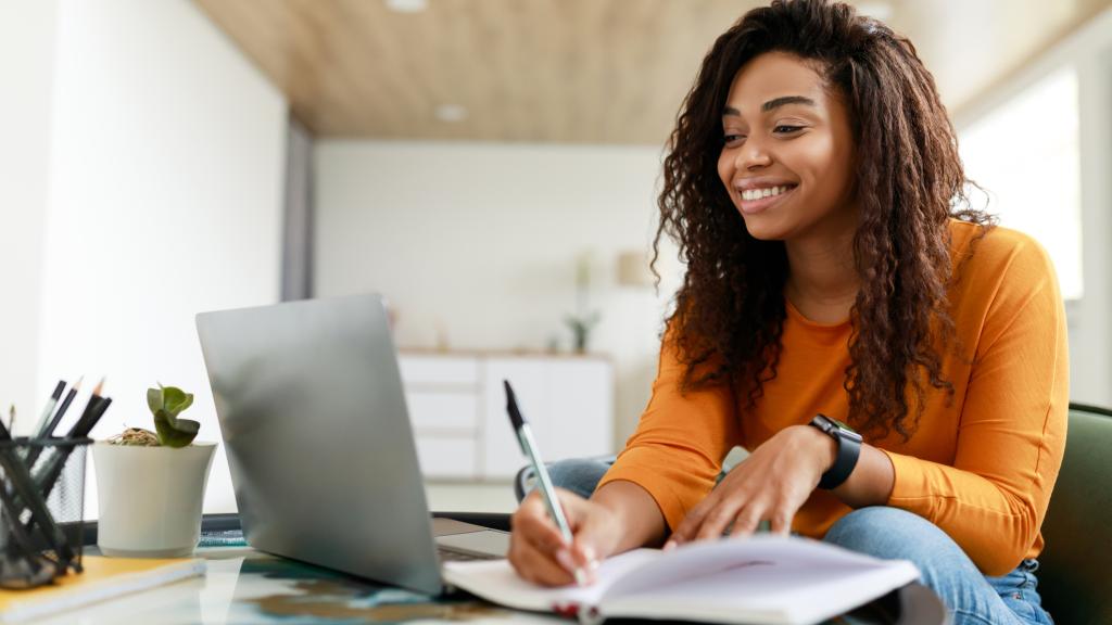 Black woman sitting at desk, using computer writing in notebook