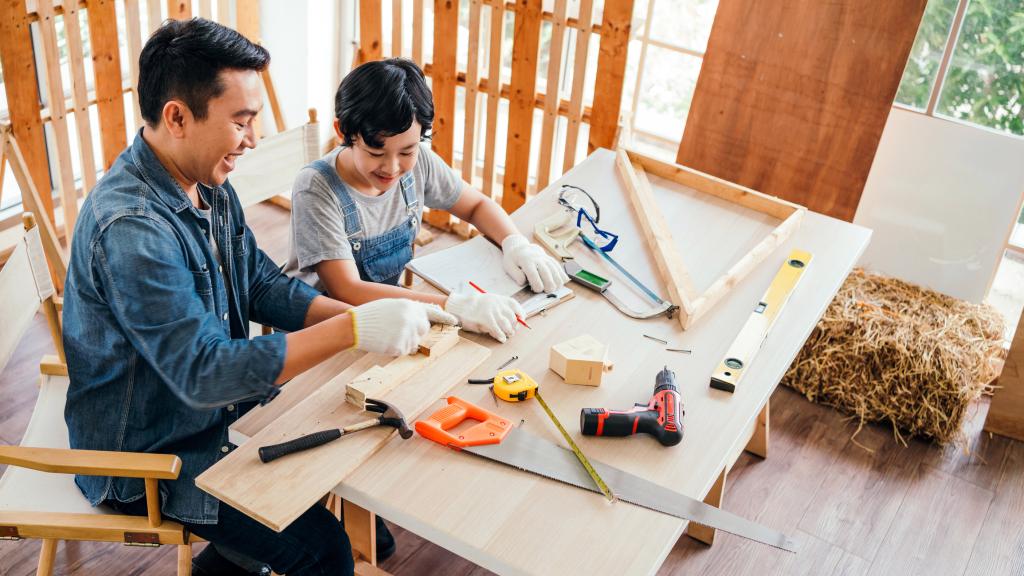 Happy Asian father and son work as a woodworker and carpenter. Father teaching his son to hammer nails on a wooden plank carefully together. carpentry working at a home workshop studio.