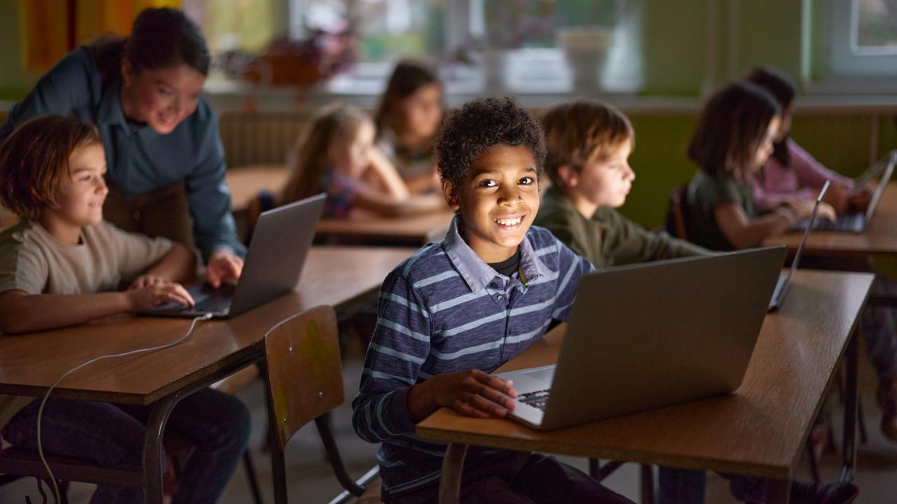 A cheerful classroom scene with elementary-aged students working on laptops. A smiling boy in the foreground looks directly at the camera, while other students and a teacher engage in learning activities in the background. The classroom is softly lit, with large windows and warm yellow curtains.