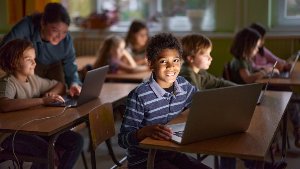 A cheerful classroom scene with elementary-aged students working on laptops. A smiling boy in the foreground looks directly at the camera, while other students and a teacher engage in learning activities in the background. The classroom is softly lit, with large windows and warm yellow curtains.