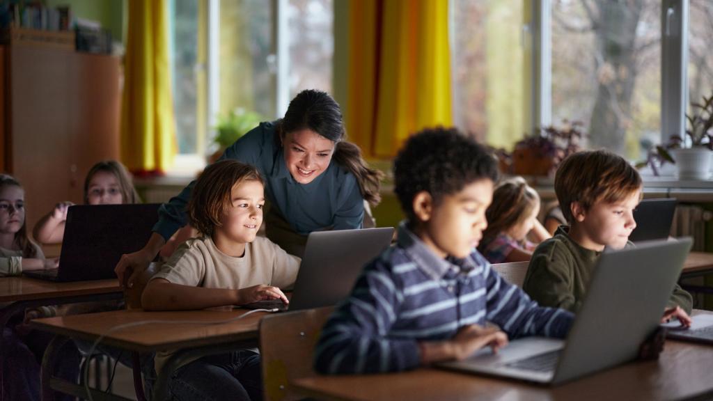 A classroom scene where a diverse group of elementary-aged students work on laptops at their desks. A smiling teacher leans over to assist one student, creating an engaging and supportive learning environment. Sunlight streams through large windows with yellow curtains in the background.