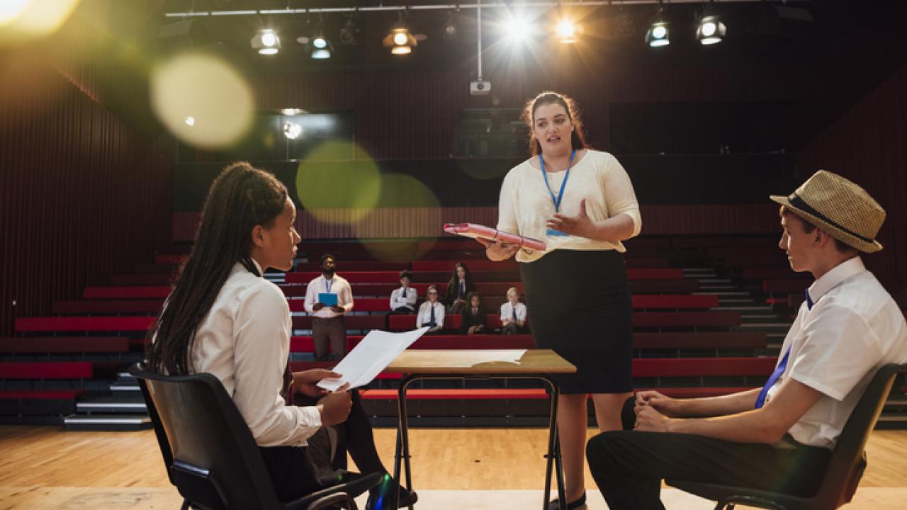 An over the shoulder view of two students who are taking notes on performace and how to improve in rehearsal. They are being watched by the rest of the drama club in the theatre.