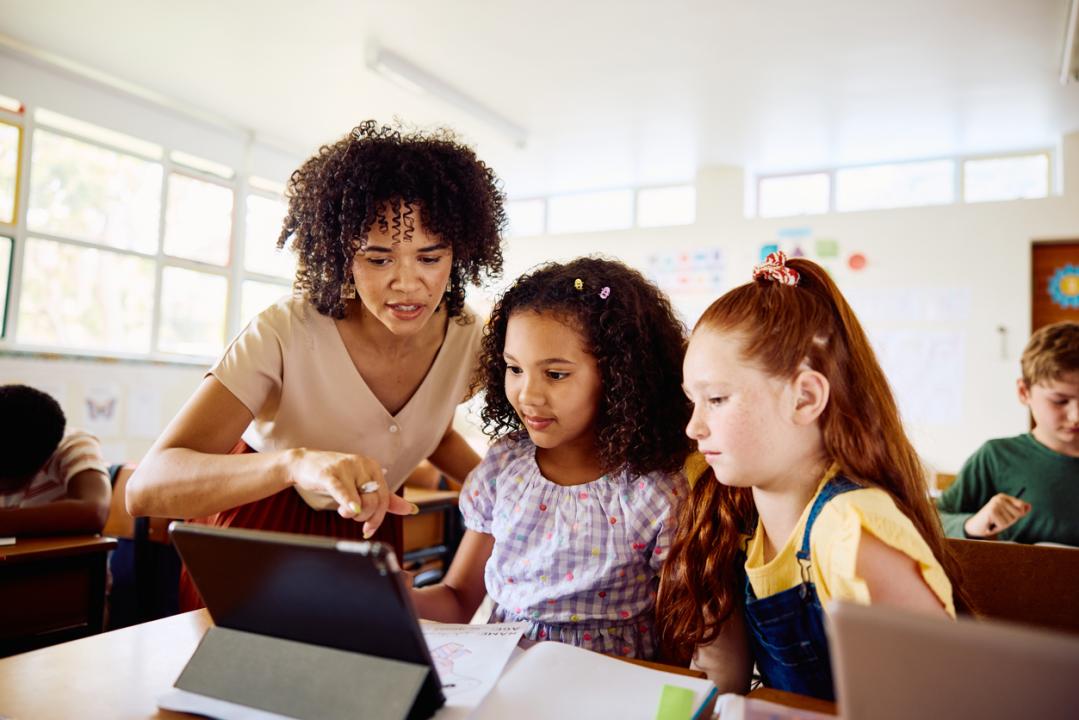 Young female teacher guiding schoolgirls using digital tablet, sitting at desk in class