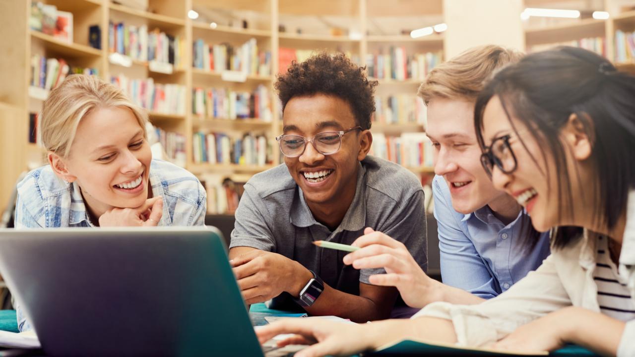 Positive excited multi-ethnic students in casual clothing lying on floor in campus library and laughing while watching curious video on laptop