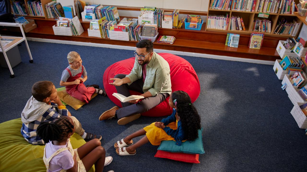 Teacher sits on a red beanbag chair reading to a small group of children seated on cushions in a library, surrounded by bookshelves.