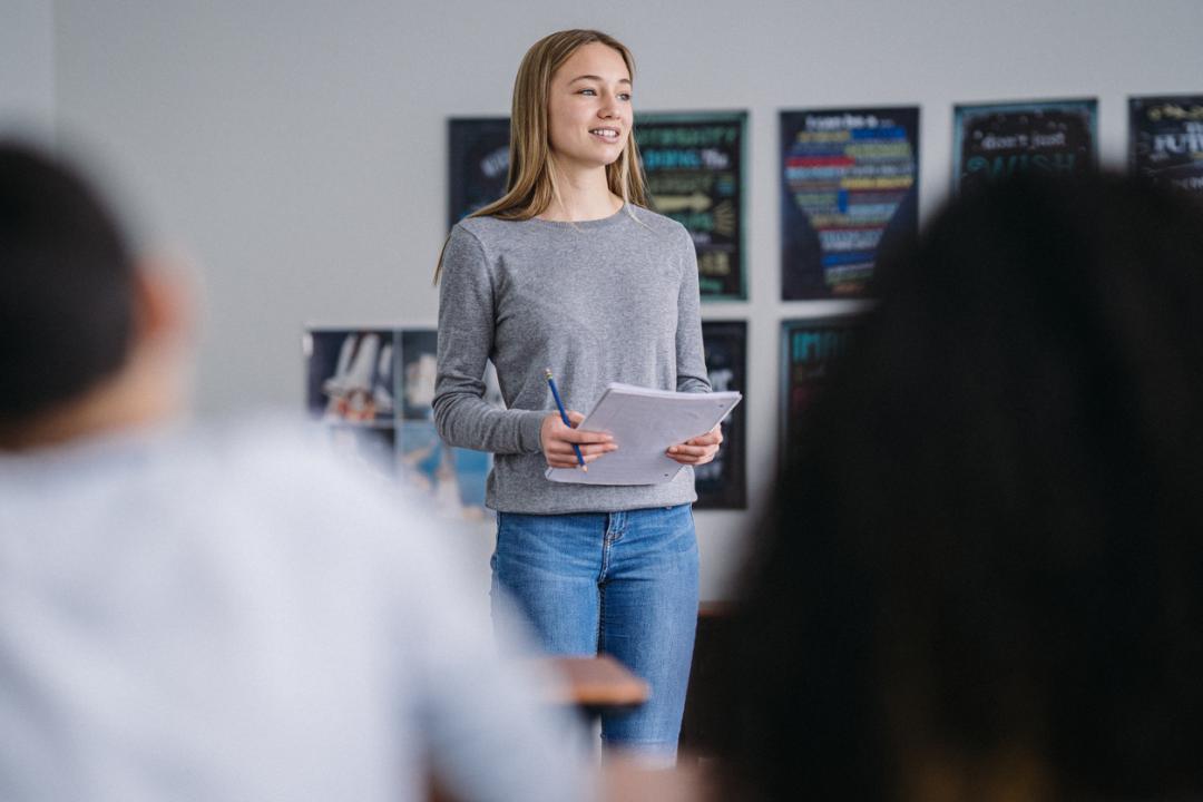 Student stands at the front of a classroom holding a notebook and pencil, smiling while presenting to classmates.