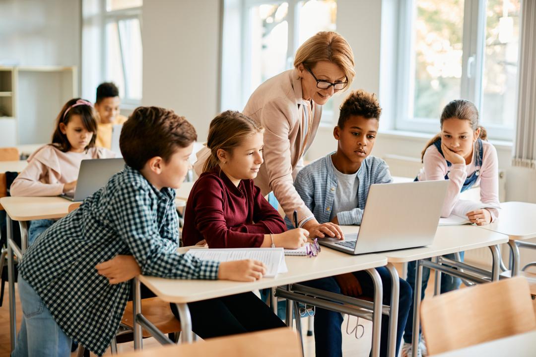 Teacher helps a group of elementary students gathered around a laptop in a classroom while other children work independently in the background.