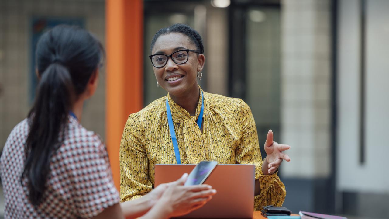Two colleagues talk at a table in a modern workspace; one woman with glasses and a lanyard gestures while speaking, while the other listens and holds a smartphone.