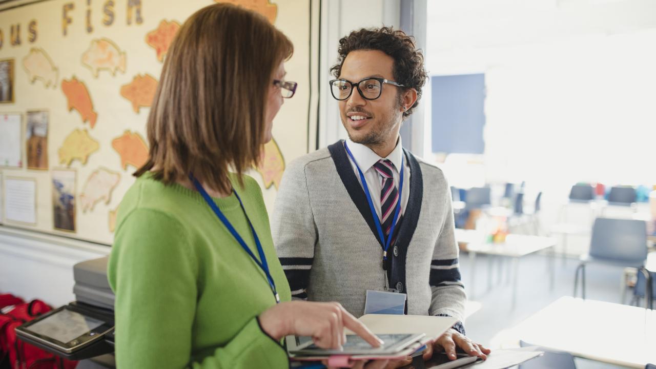 Two educators wearing lanyards stand in a classroom talking and smiling, holding tablets and folders, with student work displayed on the wall behind them.