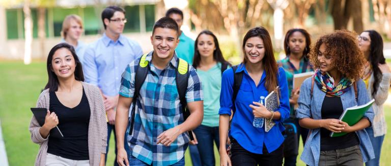 Banner size photo of diverse teenagers or young adults walking outdoors on school campus. Hispanic, Caucasian, and African American students are smiling while walking together to class. They are wearing trendy casual clothing and backpacks, and are carrying school books.