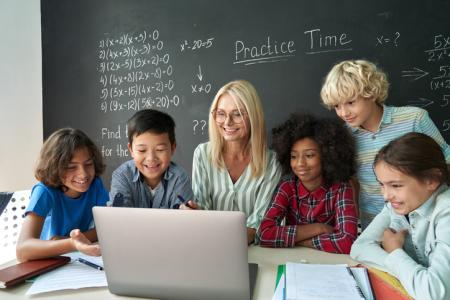 Happy diverse school children students gather at teacher table look at laptop.