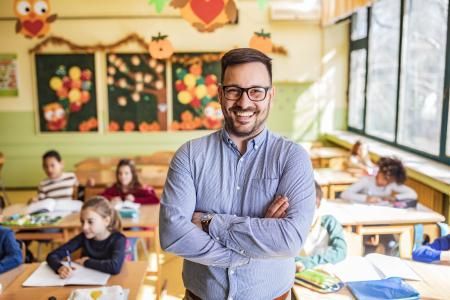 Young happy teacher standing on a class with his arms crossed and looking at camera. Elementary students are in the background.