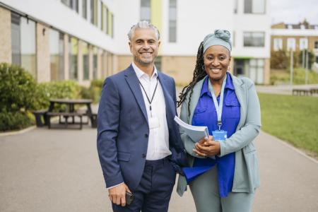 Front view of British man and woman in early 50s standing side by side and smiling at camera, education building in background.