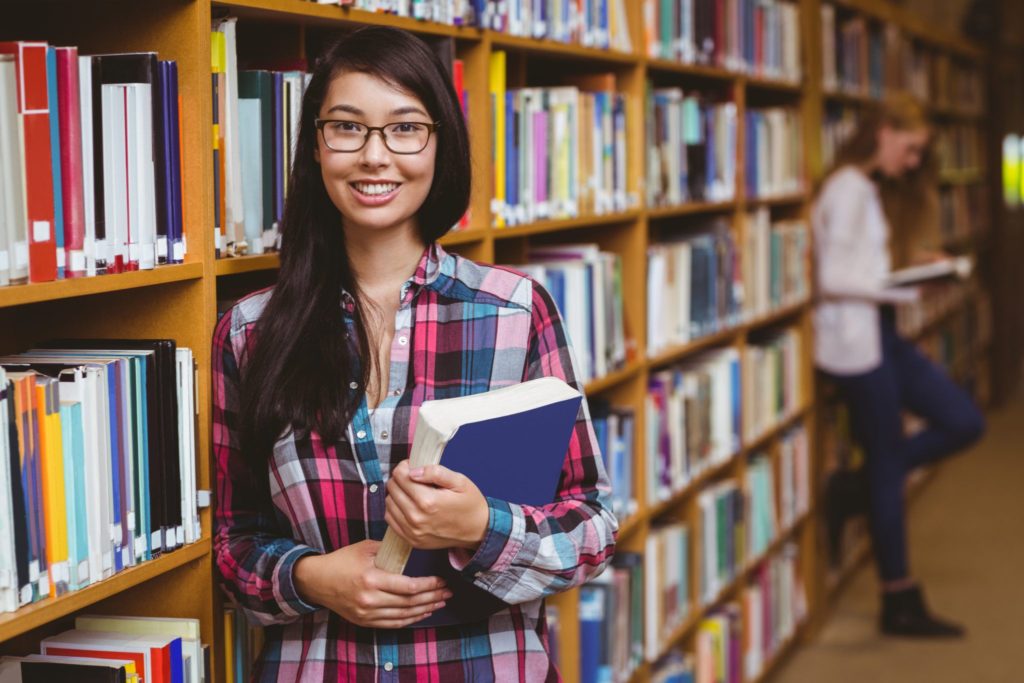 Smiling student leaning against bookshelves holding book at the university