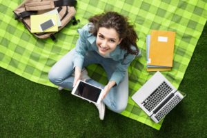 Young smiling student relaxing outdoors, she is sitting on the grass and using a laptop, summer camp concept