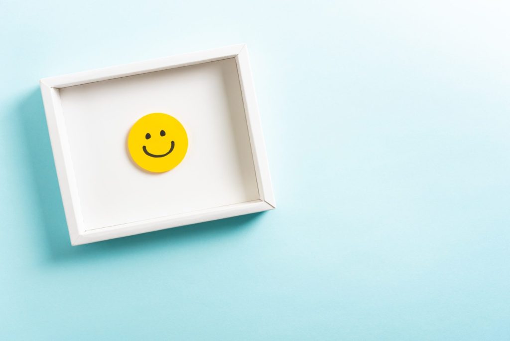 Concept of well-being, well done, feedback, employee recognition award. Happy yellow smiling emoticon face frame hanging on blue background with right empty space for text.