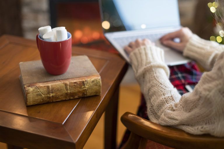 Woman hand with laptop, cup of hot cocoa and marshmallow on book on wooden table near christmas tree and fireplace.