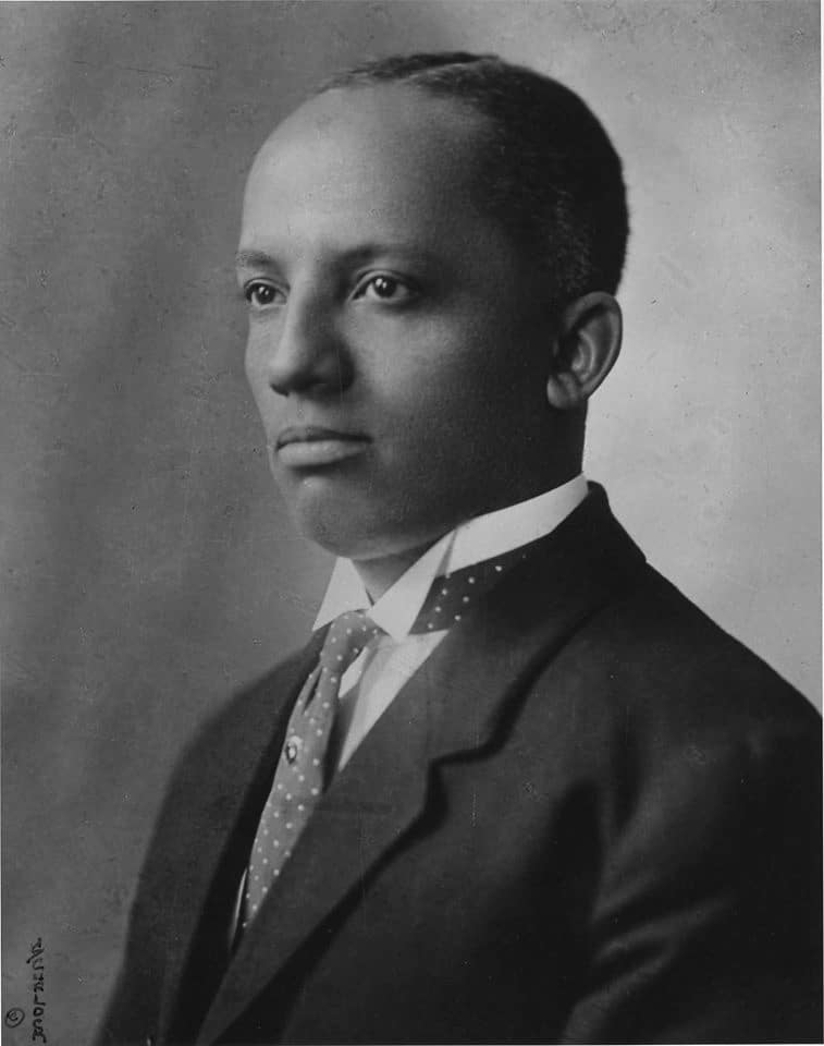 Black History Month founder Dr. Carter G. Woodson wears a tie and suit. He looks toward the left of the camera.