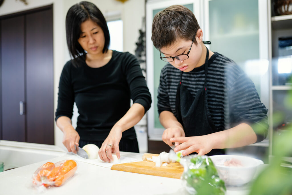 Mother watching her teenage son of differing abilities who is slicing vegetables