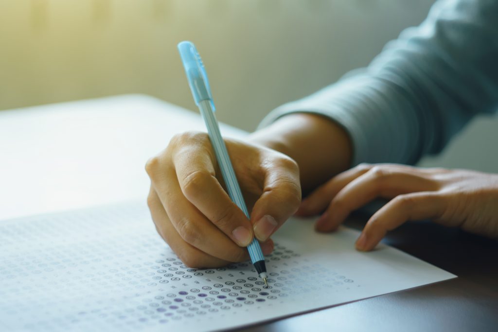 Close up of high school or university student holding a pen writing on answer sheet paper in the examination room. College students answering multiple choice questions test in the testing room in university.